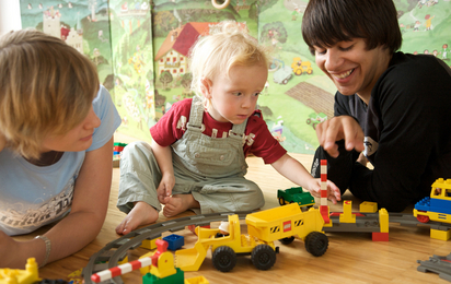 © ÖJRK/Nadja Meister Babyfit 2009. Ausbildung (Kurs) zum Babysitter. Jugendliche/Teenager. Junge und Mädchen spielen mit Kleinkind am Boden mit Legosteinen und Spielzeugeisenbahn. Querformat.